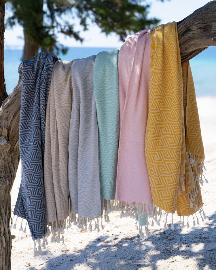 Flera tunna hammam handdukar i mintgrön, rosa, beige, grå och gul med fransar hängande på en trädgren vid strand.