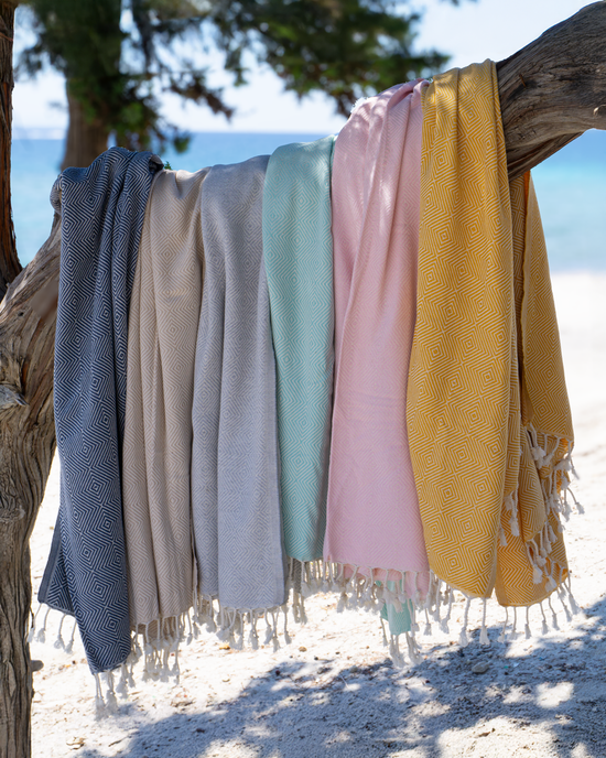 Flera turkiska hammamhanddukar i olika färger hängande på trädgren vid strand, med fransar och mjuk textur.