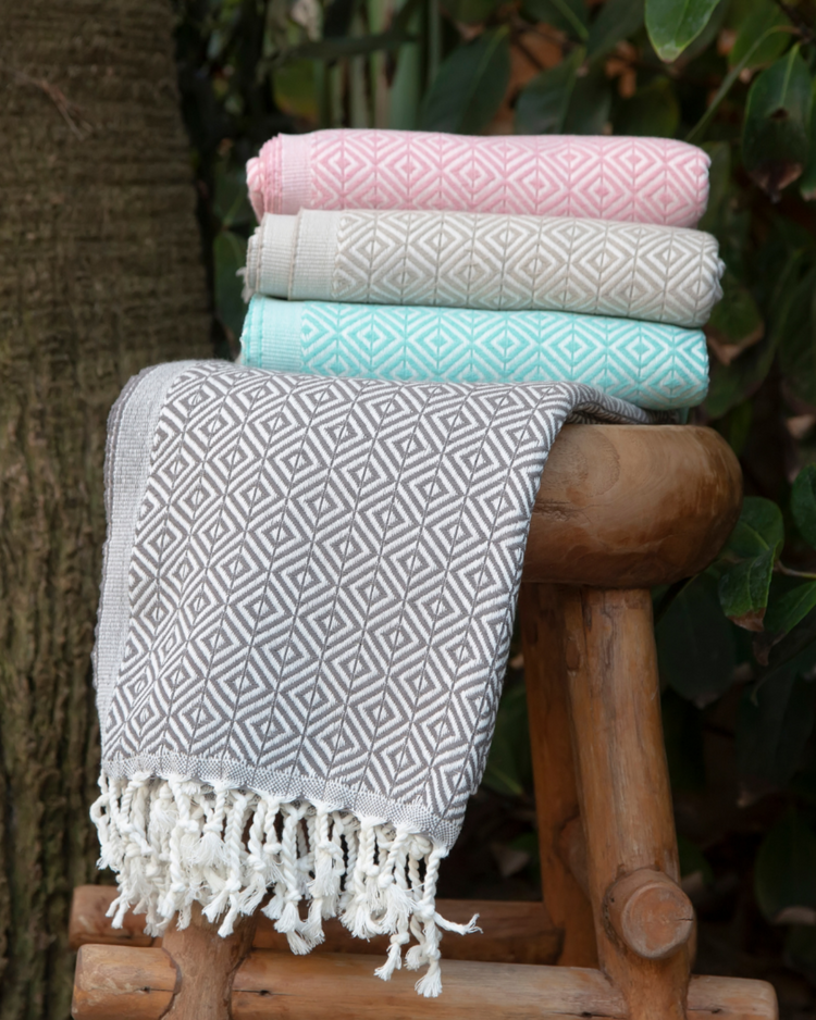 Hammam handdukar med vitmönster och fransar i grått, mint, beige och rosa staplade på träpall.