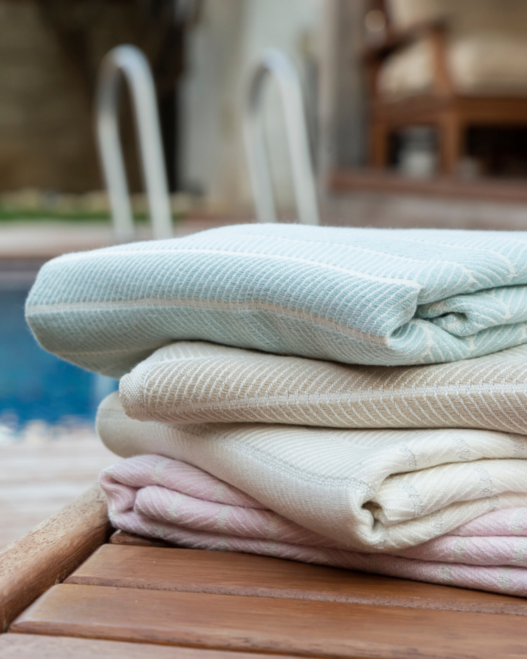 Staplad beige, rosa och ljusblå hammam-handdukar i bomull och bambu på träyta vid poolen.