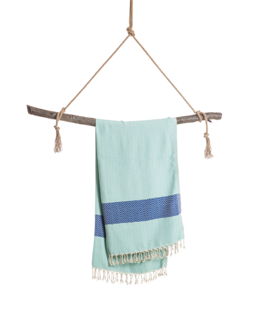 Mintblå hammamhandduk med mörkblå rand och vita fransar, hängande på rep och träpinne.