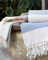 Mjuk turkisk bomullsbadhandduk med fransar i ljusblått, beige och vitt, staplade utomhus.