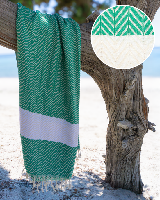 Grön och vit hammam-handduk med fiskbensmönster och fransar, hängande på trädstam.