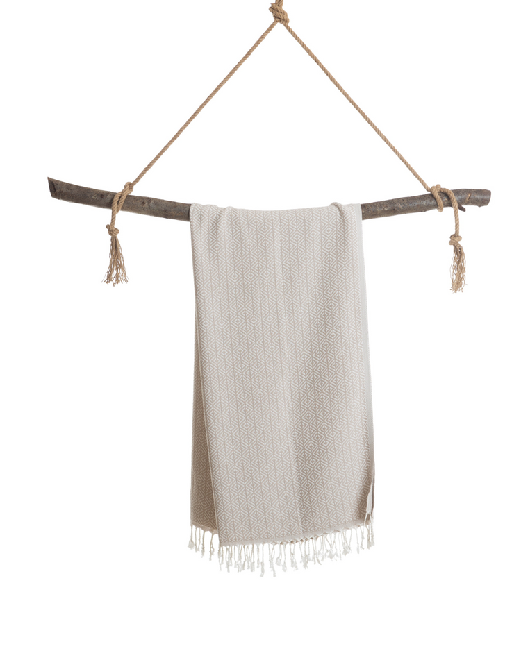 Beige hammam handduk med fransar, lätt och kompakt i oekotex bomull, hängande på trägren.