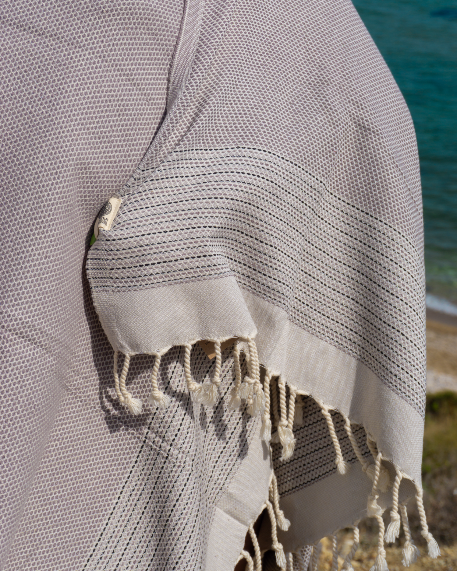 Anthracite svart turkisk hammam handduk med fransar och finmaskigt mönster, hängande vid strandmiljö.